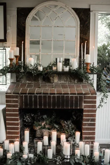 A brick fireplace decorated with greenery, numerous white candles of varying heights, and a large arched mirror above the mantel, creating a cozy and elegant atmosphere.