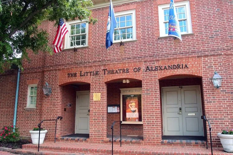 The front entrance of The Little Theatre of Alexandria, a red brick building with three flags above, two gray doors, potted plants, and a poster display under an arched entryway.