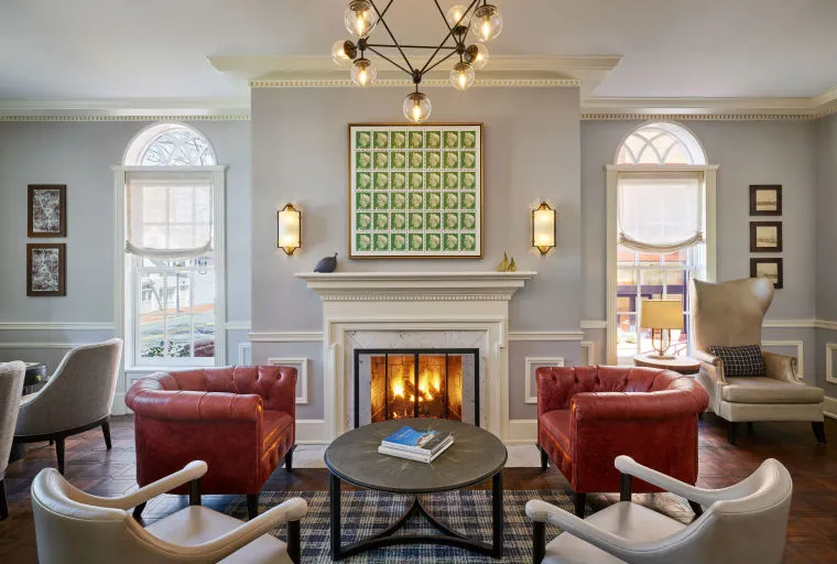 A cozy, elegant living room with a lit fireplace, two red leather chairs, neutral-toned armchairs, a round coffee table, and arched windows letting in natural light. Artwork and lamps decorate the light gray walls.