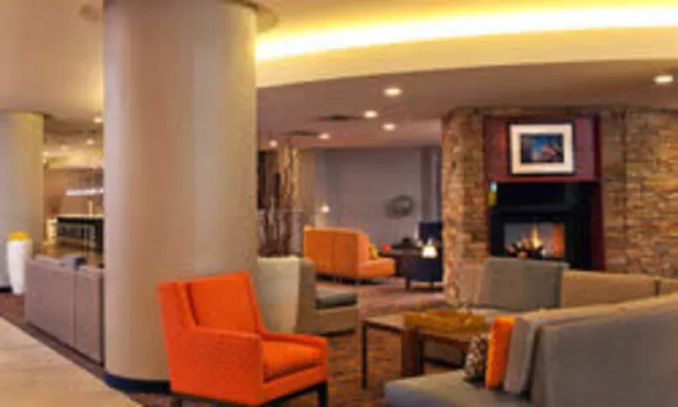 Modern hotel lobby with contemporary seating, orange and gray chairs, a stone fireplace, soft lighting, and framed artwork on the wall, creating a warm and inviting atmosphere.