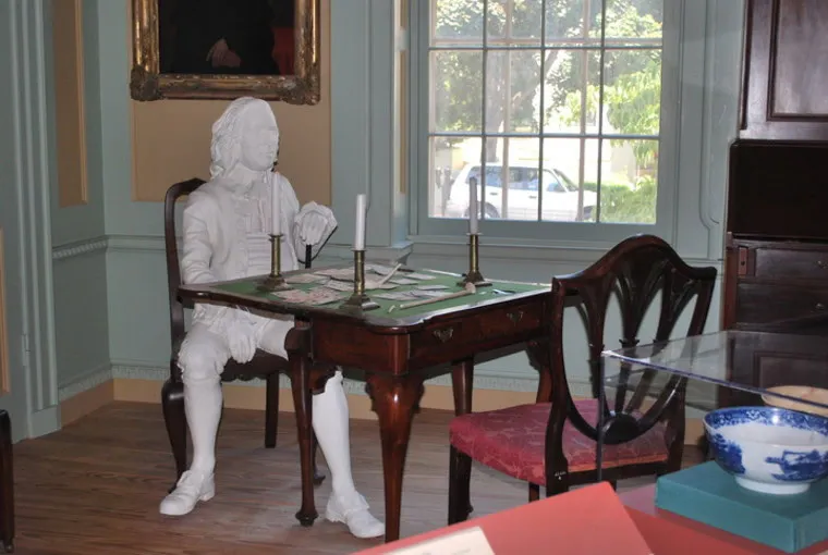 A white statue of a seated man in historical clothing sits at a wooden table with candlesticks, papers, and a chair nearby in a museum room with a large window and framed pictures.