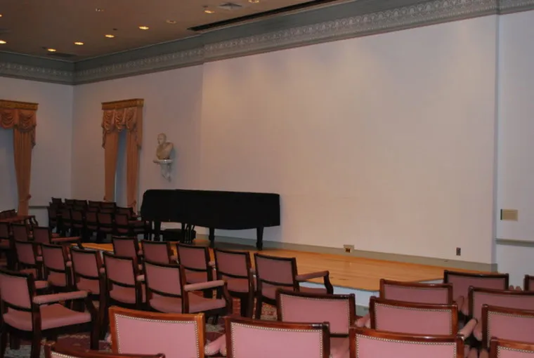 Rows of empty pink-cushioned chairs face a small stage with a black piano and a white bust on the left wall; the room has ornate trim and tall curtained windows.