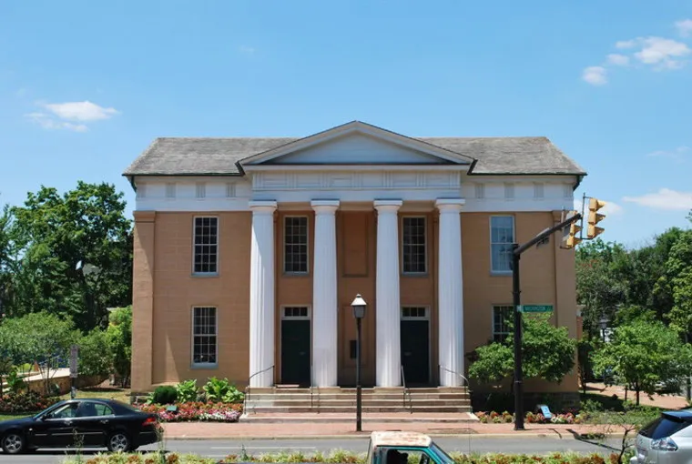 A large tan brick building with four white columns, tall windows, and double doors sits on a sunny day. Several cars are parked in front, and trees and plants surround the building.