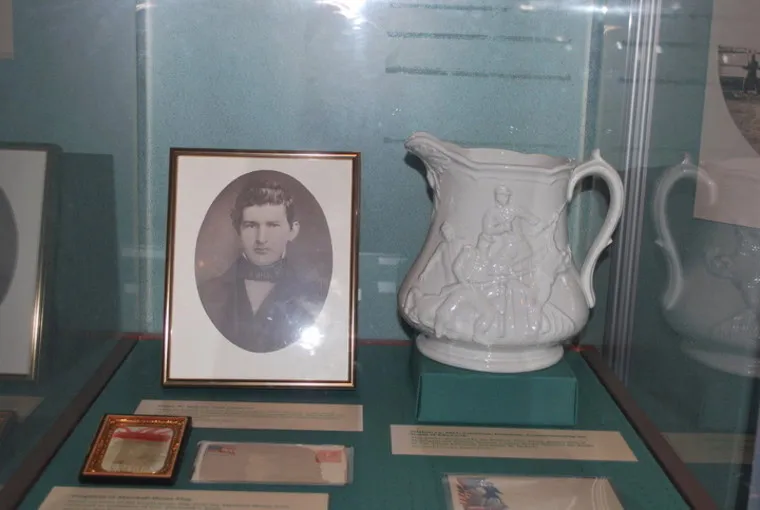 A framed black-and-white portrait of a man in formal attire is displayed next to a large ornate white pitcher with embossed figures, all set in a glass museum case.