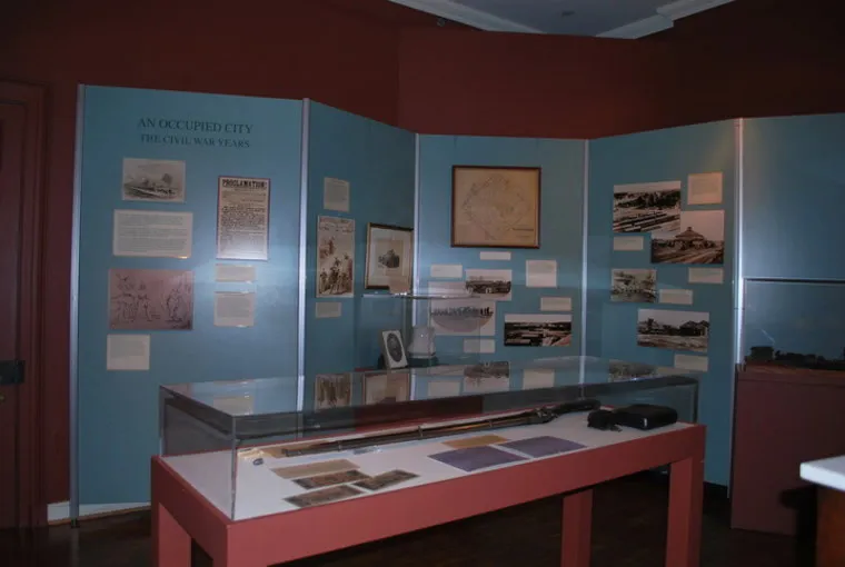 A museum exhibit about the Civil War features historical photos, documents, and maps on blue display boards, with a glass case in front containing artifacts including a rifle and papers.