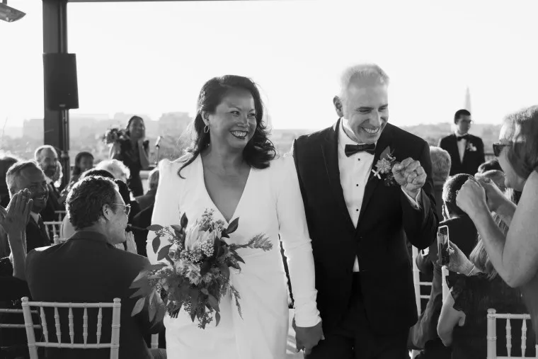 A newlywed couple walks down the aisle, smiling and holding hands, as guests applaud. The bride wears a white dress and holds a bouquet; the groom is in a suit. The setting appears bright and joyful.
