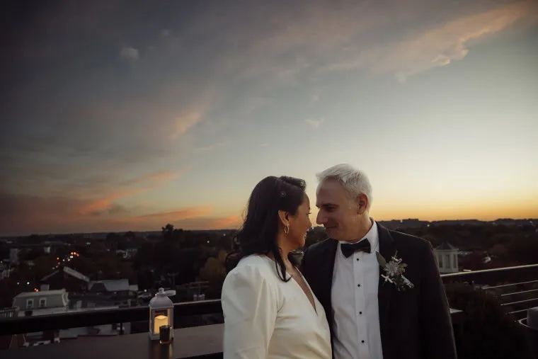 A couple dressed in formal wedding attire stands close together on a rooftop at sunset, with warm, soft lighting and a lantern on the ledge beside them. The sky is painted with clouds and orange hues in the background.