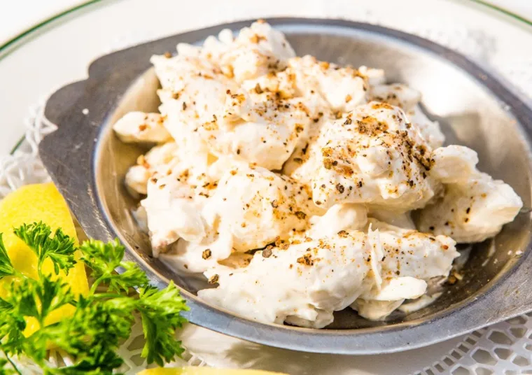 A metal dish filled with creamy crab meat, topped with cracked black pepper, sits on a white plate. Fresh parsley and lemon wedges are garnished on the side.