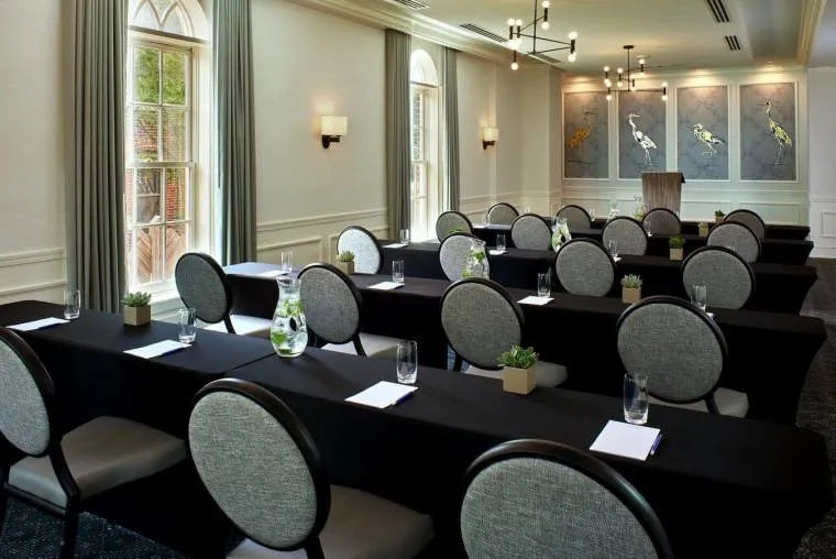 A well-lit conference room with three rows of black-covered tables, gray chairs, notepads, glasses, water pitchers, and small potted plants facing a podium, with large windows and decorative wall art.