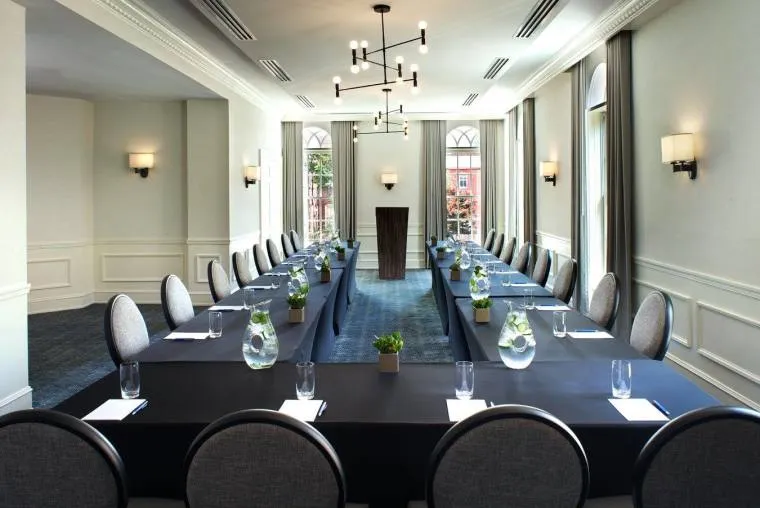 A modern, well-lit conference room with two long tables arranged in a U-shape, set with glasses, notepads, water pitchers, and small potted plants. A wooden podium stands at the front, and large windows let in natural light.