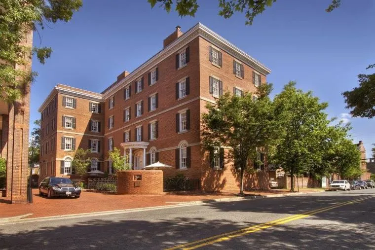 A large, four-story brick building with white trim and black shutters, surrounded by trees. A car is parked outside on a red brick driveway next to a quiet street under a clear blue sky.