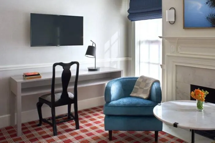 A modern, bright room with a wall-mounted TV above a minimalist desk and chair, a blue armchair with a pillow, a round marble table with orange flowers, and a window with a blue shade near a white fireplace.