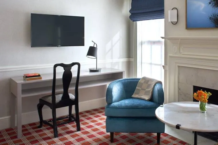 A modern, bright room with a wall-mounted TV above a minimalist desk and chair, a blue armchair with a pillow, a round marble table with orange flowers, and a window with a blue shade near a white fireplace.