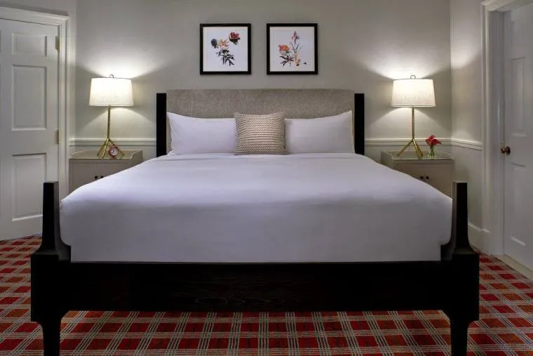 A neatly made bed with white linens is centered between two nightstands, each with a lamp. Two framed flower prints hang on the wall above the bed. The room has patterned red carpet and neutral walls.