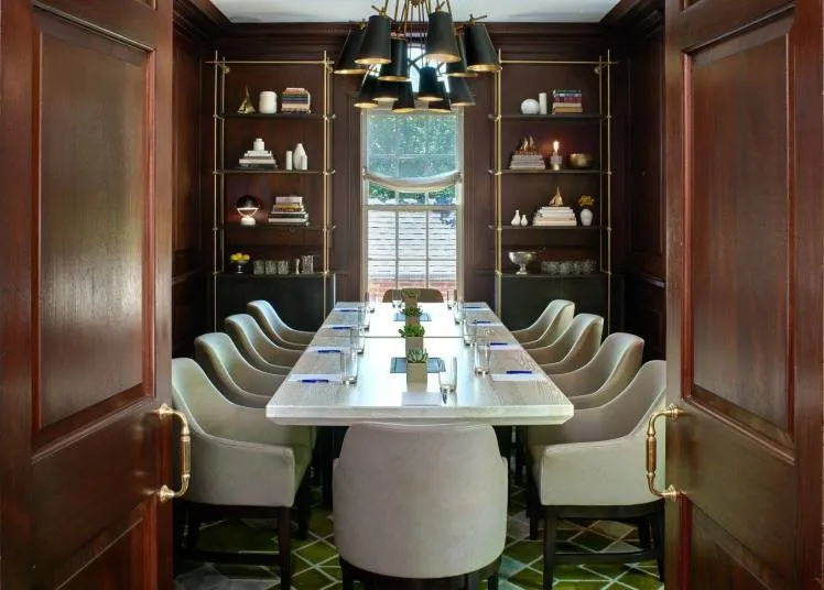 A modern conference room with a long marble table, twelve upholstered chairs, dark wood paneling, built-in shelves with decor items, and a contemporary black chandelier, viewed through open double doors.