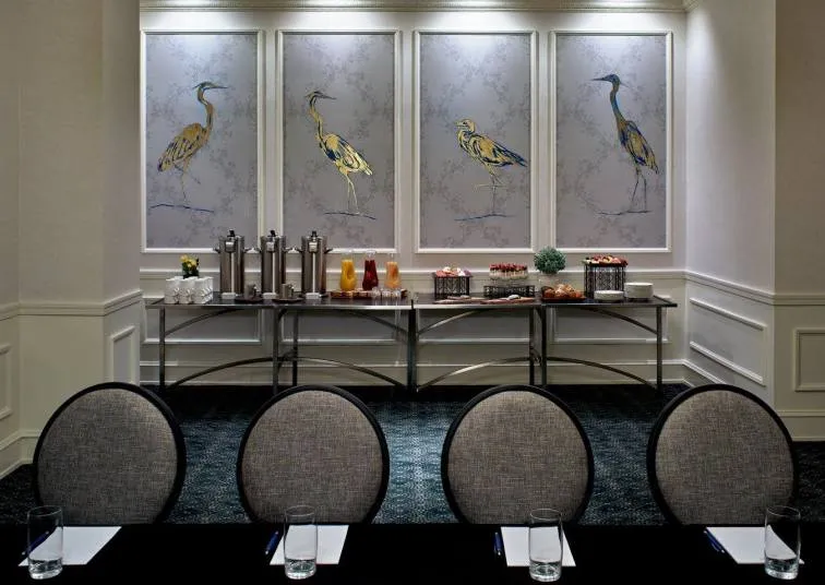 A conference room setup with four chairs facing a table, glasses and notepads, and a refreshment station behind them with drinks and snacks. Four large bird illustrations hang on the wall above the refreshments.