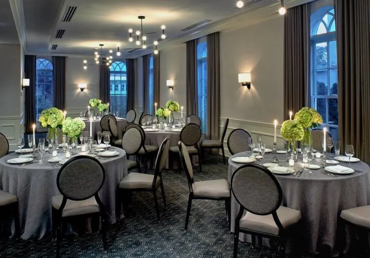 Elegant dining room with round tables covered in gray tablecloths, set for a formal event. Each table has green flower centerpieces and candles. Large windows with drapes line the walls, and modern pendant lights hang from the ceiling.