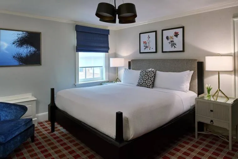 A modern hotel room with a large bed, white linens, two nightstands with lamps, framed floral art above the bed, a blue armchair, plaid carpet, and a window with a blue shade.