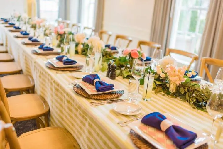 A long, elegantly set dining table with striped tablecloth, floral centerpieces, wooden chairs, and place settings featuring woven chargers, plates, glasses, and blue napkins tied with ribbon. Natural light from nearby windows.