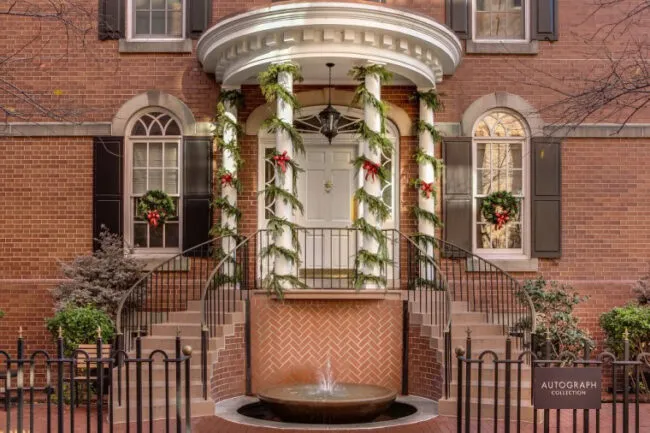 A brick building entrance decorated with holiday garlands and wreaths with red bows. There’s a white door, curved double staircase, a small fountain in front, and a sign that reads “Autograph Collection.”.