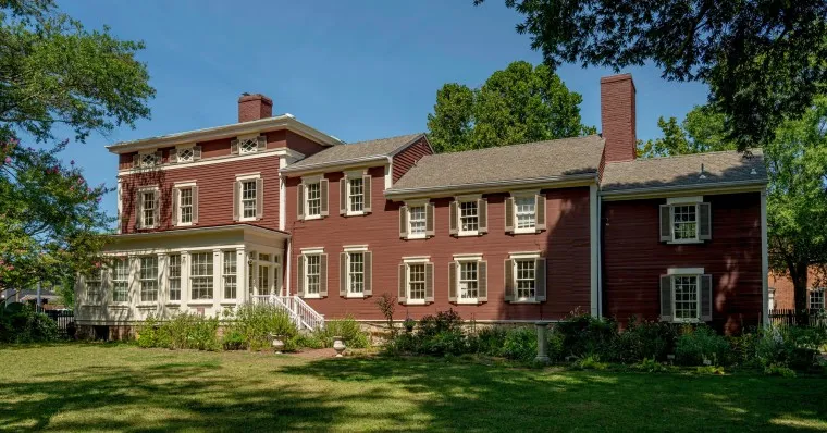 A large historic red wooden house with white trim, multiple chimneys, many windows, and a sunroom, surrounded by green grass, trees, and shrubs under a clear blue sky.