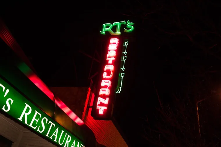 A neon sign at night reads RT’s RESTAURANT in green and red letters. The sign is mounted on a building, with part of the restaurant’s exterior visible, lit by green and red lights.