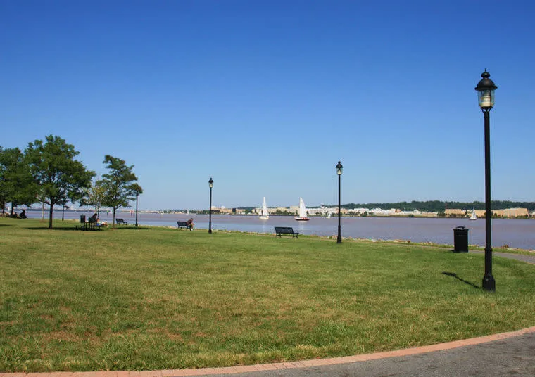 A grassy park with benches and lampposts overlooks a calm river with sailboats. Trees line the left edge, and buildings are visible across the water under a clear blue sky.