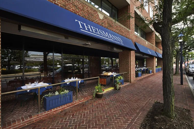 Street view of Theismann’s restaurant with blue awnings and outdoor seating along a brick sidewalk, with tables set and potted plants lining the patio area.