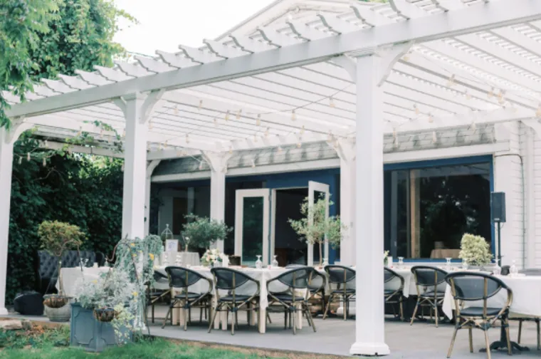 Outdoor dining area under a white pergola with hanging string lights, several round tables with chairs set for a meal, surrounded by greenery and potted plants next to a white building with large windows.