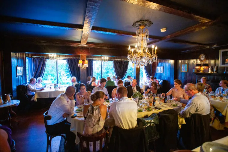 A group of people dressed formally sit around tables in a dimly lit restaurant with chandeliers, eating and conversing. The room has dark wood walls, large windows with curtains, and a cozy, elegant atmosphere.