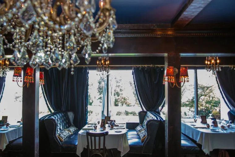 Elegant restaurant interior with dark velvet booths, crystal chandeliers, and tables set with white linens near large windows, letting in natural light.