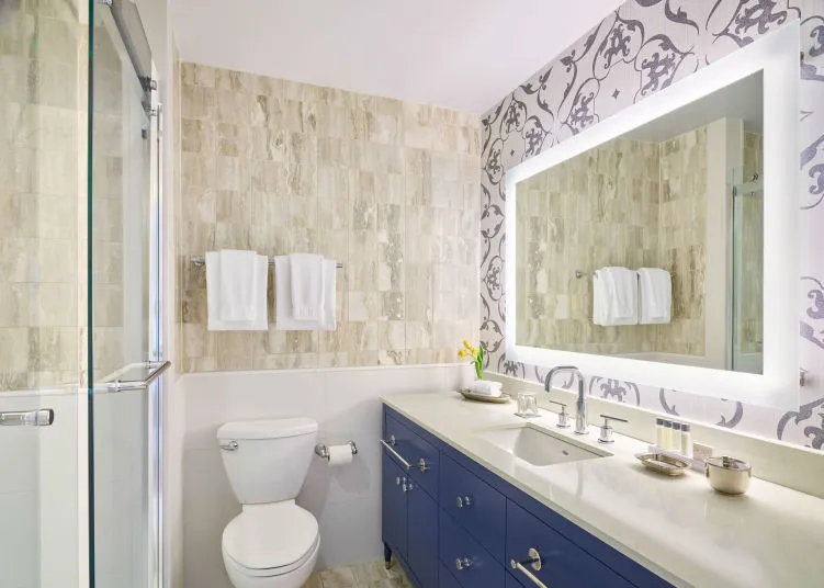 Modern bathroom with a large backlit mirror, blue vanity with a double sink, white countertop, wall-mounted towels, toilet, glass shower, and decorative tile and wallpaper accents. A vase with yellow flowers sits on the counter.