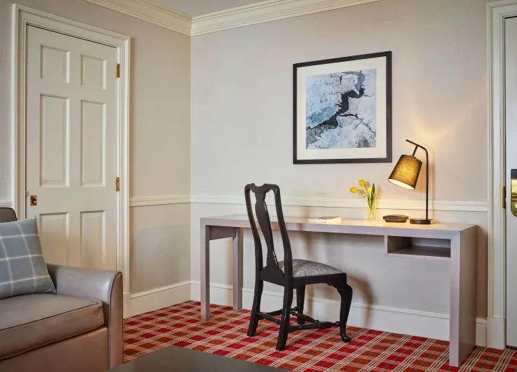 A tidy hotel room desk setup with a black chair, desk lamp, yellow tulips in a vase, notepad, abstract wall art above, cream walls, and red plaid carpet. Part of a beige sofa and white door are visible.
