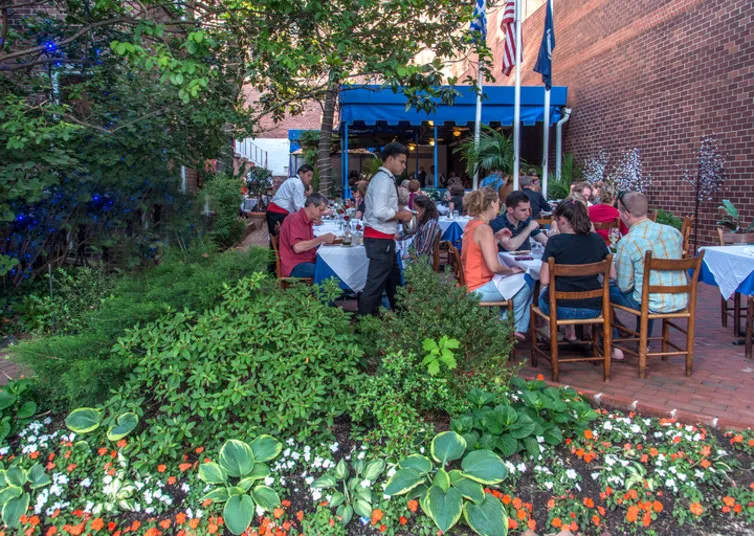 People dine at outdoor tables in a garden patio restaurant, surrounded by flowers and greenery. A waiter serves one table. Blue awnings and American flags are visible near the brick building in the background.