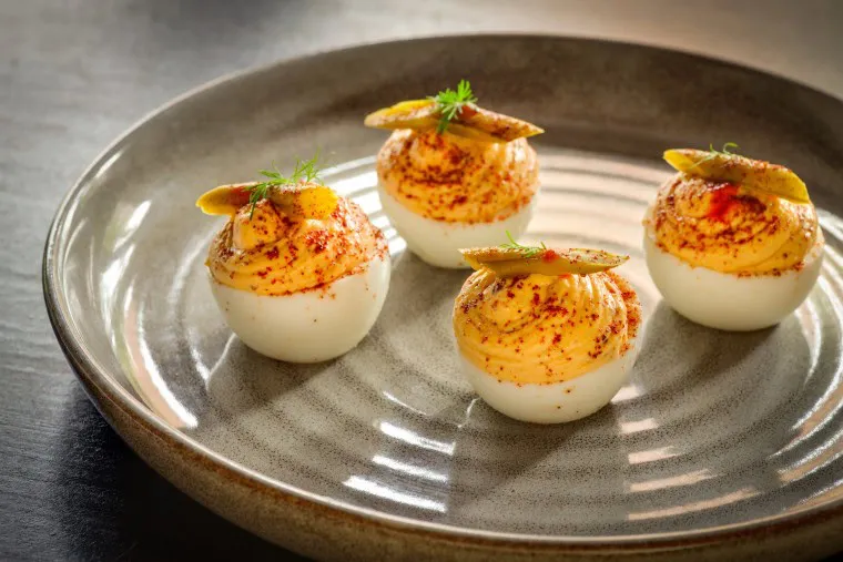 Four deviled eggs are arranged on a striped ceramic plate. Each egg half is topped with creamy filling, a slice of pickle, a sprinkle of paprika, and a small herb garnish. The plate is set on a dark surface.
