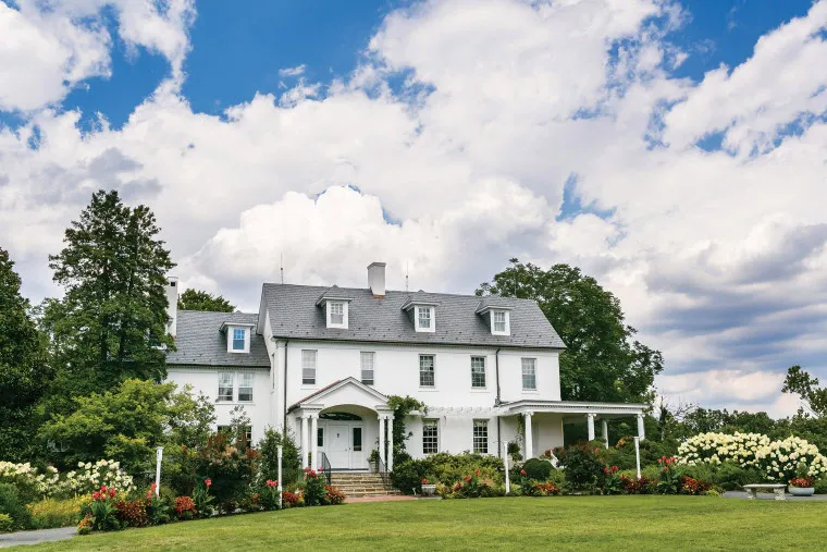 A large white colonial-style house with dark gray roof, surrounded by lush green lawn, colorful flowers, and trees, set under a bright blue sky with scattered clouds.