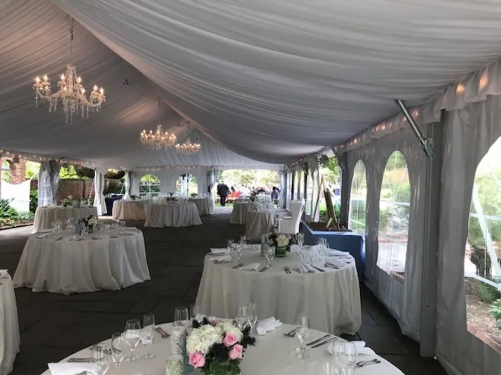 A white tent with chandeliers and draped ceiling is set up for an event, featuring round tables with white tablecloths, elegant place settings, and floral centerpieces. Natural light enters through clear side panels.