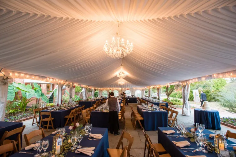 A large white tent with chandeliers and draped ceiling is set up for an elegant event, featuring long tables covered in navy blue tablecloths, wooden chairs, and glassware, with greenery visible outside.