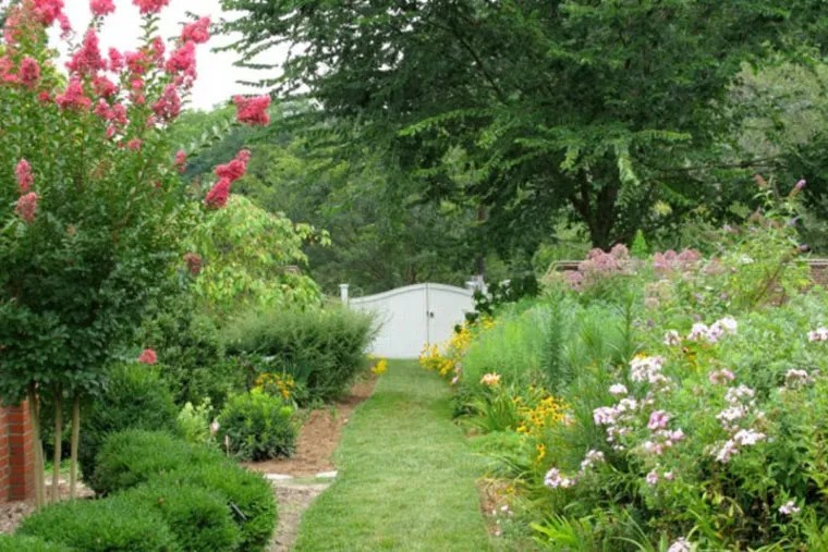 A lush garden path lined with vibrant flowering plants and bushes leads to a closed white gate, surrounded by tall green trees and colorful blossoms.
