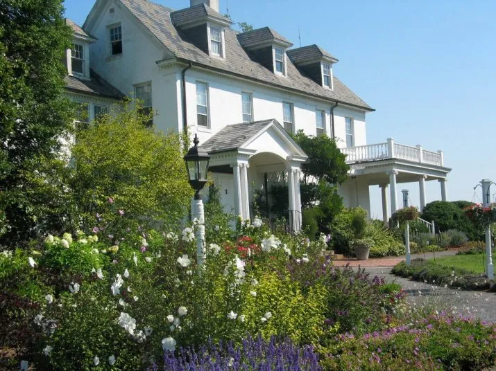 A large, white, three-story house with dormer windows and a wraparound porch is surrounded by a colorful garden of blooming flowers and green shrubs under a clear blue sky.