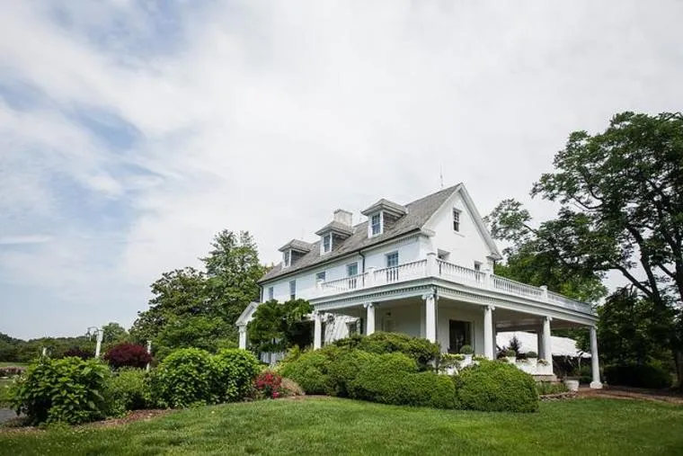 A large white two-story house with a wraparound porch, surrounded by green bushes, trees, and a well-kept lawn under a partly cloudy sky.