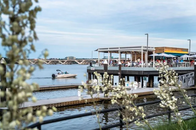 People dine at an outdoor waterfront restaurant while a motorboat passes by on the river. Flowering plants are in the foreground and a long bridge is visible in the distance under a partly cloudy sky.