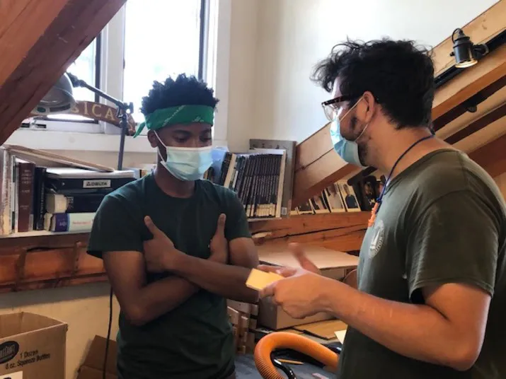 Two people wearing face masks talk in a room with bookshelves, a window, and wooden beams. One person has a green bandana and crossed arms, while the other holds a piece of paper and gestures.