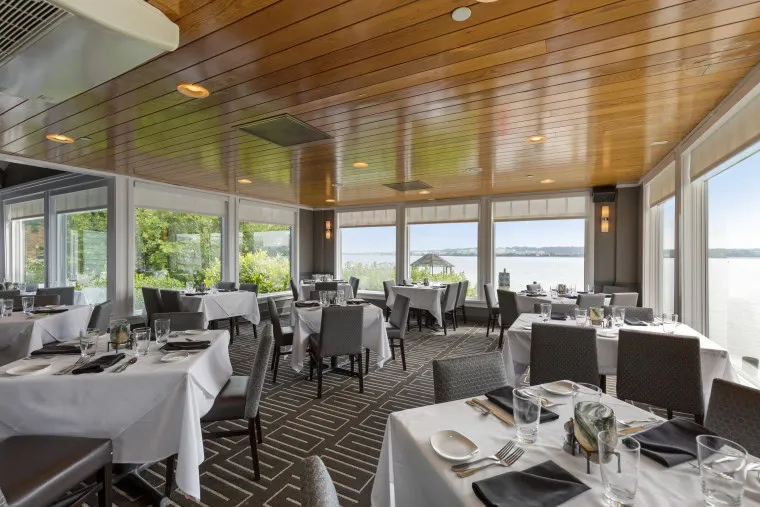 A bright, elegant restaurant dining room with large windows overlooking a lake, tables set with white cloths, glassware, and silverware, and upholstered chairs on a geometric-patterned carpet.