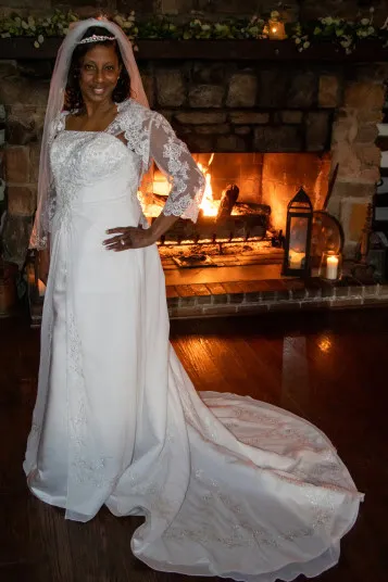 A smiling woman in a white wedding dress and veil stands indoors in front of a stone fireplace with a burning fire and candles, her hand on her hip and the train of her dress spread behind her.