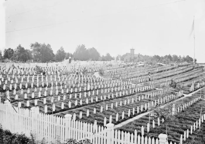 Rows of white headstones fill a cemetery enclosed by a white picket fence, with trees and a distant building visible in the background under an overcast sky.