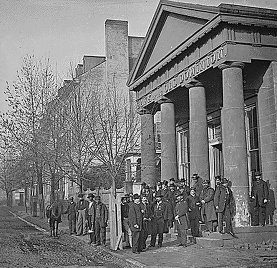 Black-and-white photo of a group of men in uniforms standing outside a large columned building labeled Old Dominion, with trees and a horse along the street.