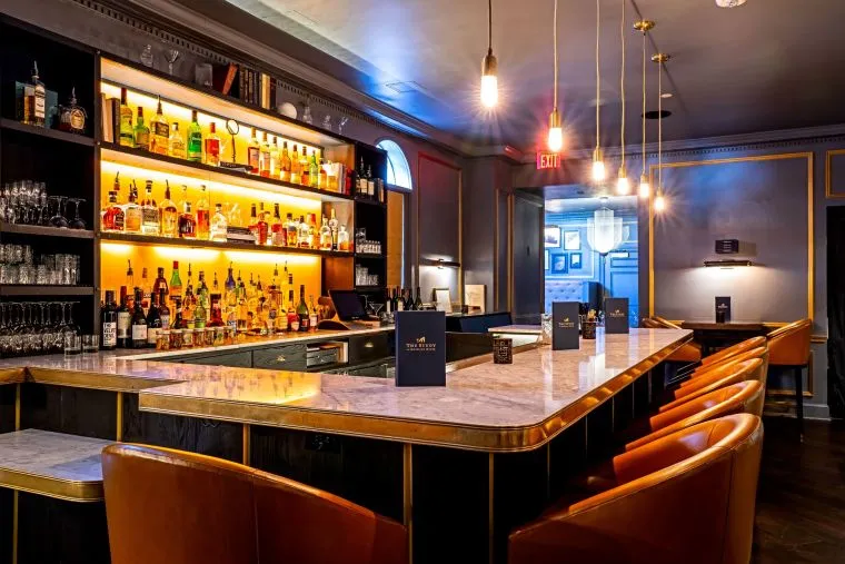 A stylish, modern bar with a marble counter, tan leather barstools, illuminated shelves displaying liquor bottles, hanging pendant lights, and dark walls with gold trim and decorative accents.