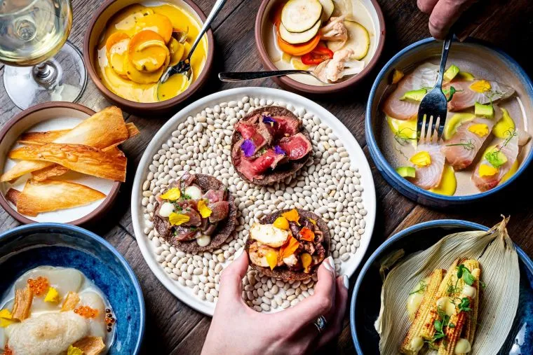 A colorful spread of gourmet dishes on a wooden table, featuring small plates of ceviche, root vegetables, grilled corn, and three tostadas on beans, with two hands reaching for food.