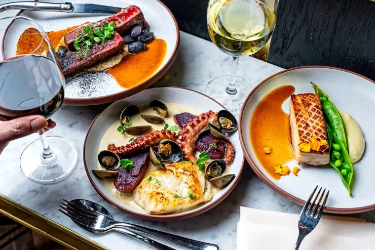 Three elegant plates of food on a marble table: grilled steak, seafood with octopus and clams, chicken with pea and mash, plus glasses of white and red wine; a hand holds a wine glass.